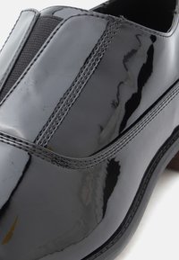 Μαύρο παπούτσι από λουστρίνι με ελαστικές πλαϊνές επιφάνειες, χαρακτηρίζεται από λεία, λαμπερή επιφάνεια και διακριτικές λεπτομέρειες ραφής κατά μήκος των ραφών.