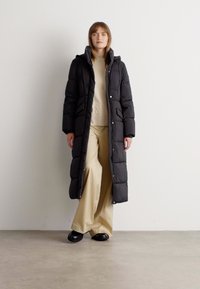 Νεαρή γυναίκα που φοράει μαύρο χοντρό παλτό πάνω από μπεζ πουλόβερ και φαρδιά μπεζ παντελόνια, στέκεται σε ομαλή επιφάνεια μπροστά σε λευκό τοίχο.