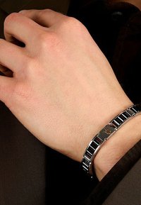 Svart och silver metallarmband med växlade rektangulära länkar och en central logodetalj, bärs på en ljus hudfärgad hand.