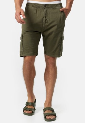 Mann trägt olivgrüne Cargo-Shorts und passende grüne Sandalen mit Schnallen, steht mit Händen in der Nähe der Taschen vor weißem Hintergrund.