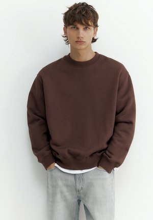 Jeune homme aux cheveux bruns ébouriffés portant un sweat-shirt marron foncé oversize et un jean gris clair, debout devant un fond blanc uni.