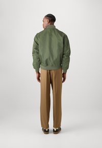Jachetă bomber verde cu o textură netedă, cu manșete și tiv ribbate. Asortată cu pantaloni maro, evidențiind o fixare lejeră și un design ajustat.