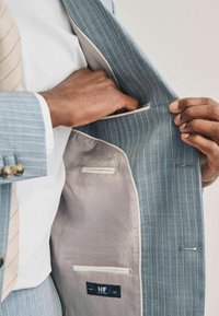 Veste de costume bleu clair à rayures avec une doublure lisse et à motif. Comprend des poches intérieures et une étiquette cousue en noir et bleu.