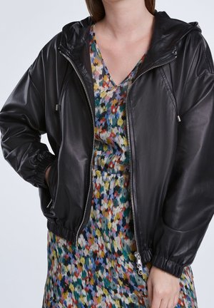 Person wearing a black zippered leather jacket over a multicolored abstract patterned dress, with one hand in pocket and the other resting by side.