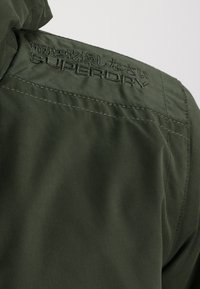 Mørkegrøn jakke med en glat tekstur, der har syet detaljer og logoet "SUPERDRY" broderet på skulderen.