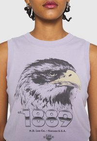 Vaaleanlila hihaton T-paita, jossa suuri mustavalkoinen kotkapainatus. Alla tekstit "est. 1889" ja "H.D. Lee Co. - Kansas U.S.A.".