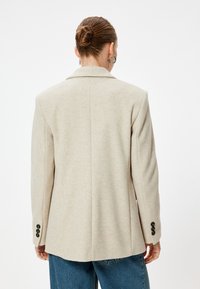 Blazer beige con un design strutturato, colletto a scavo e tre bottoni neri su ciascun polsino. Realizzato in un tessuto morbido e texturizzato.