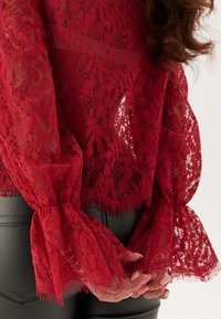 Rød blondebluse med lange, klokkeformede ærmer, der har frynset kant. Stoffet er semi-gennemsigtigt, hvilket afslører en glat sort baggrund nedenunder.