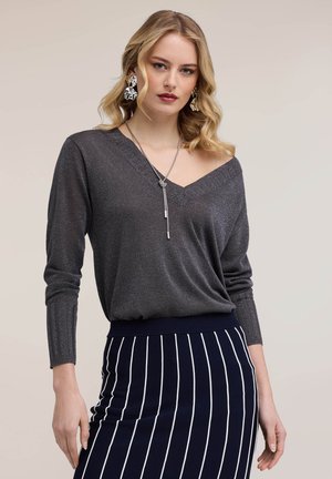 Kobieta o falowanych blond włosach, ubrana w błyszczącą ciemnoszarą bluzkę z odkrytymi ramionami, srebrny naszyjnik, wyróżniające się kolczyki oraz granatową spódnicę z białymi pionowymi paskami.