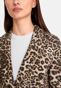 Leopard print blazer with sharp lapels, featuring a textured fabric. Worn over a light gray, ribbed shirt. Close-up view of the neckline.