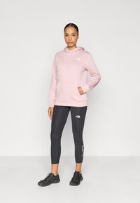 Rózsaszín kapucnis pulóver elülső zsebbel, fekete leggingsszel, amelyen logó látható. A modell fekete sportcipőt visel. Simább szövet.