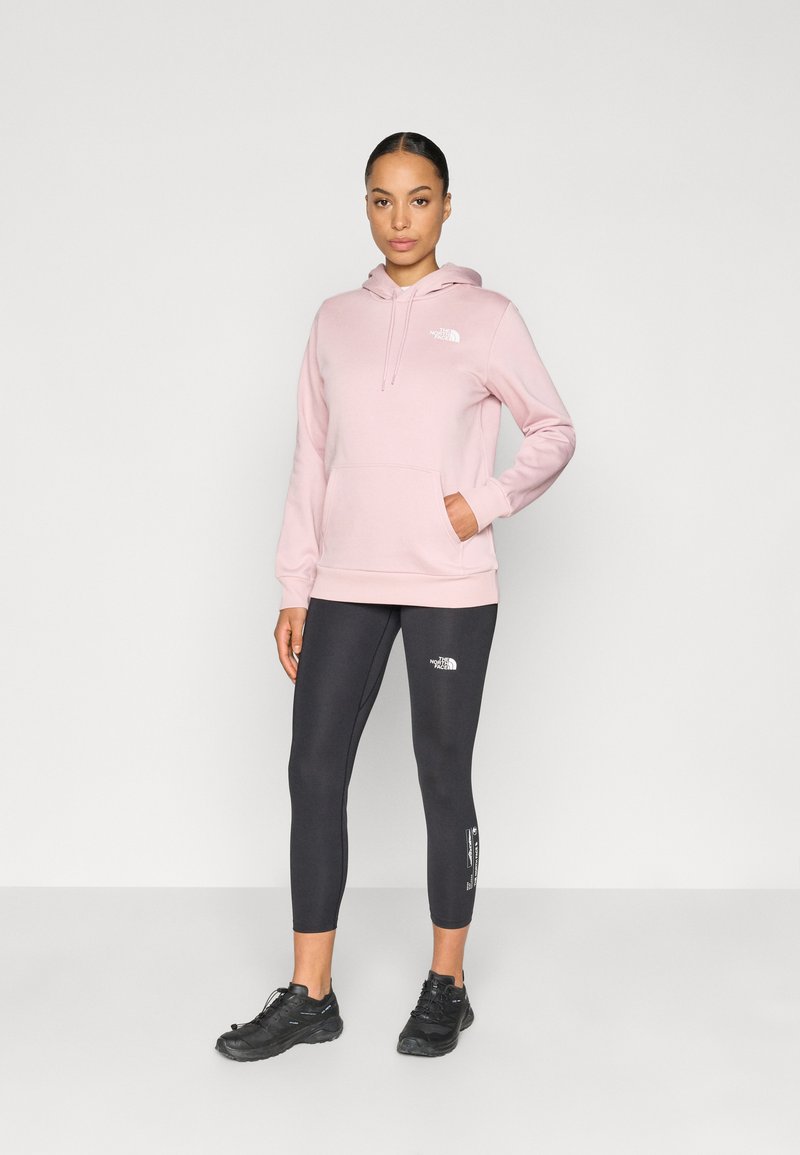 Lyserød hættetrøje med forlomme, kombineret med sorte leggings med logo. Modellen har sorte sports sko på. Glat stoftekstur.