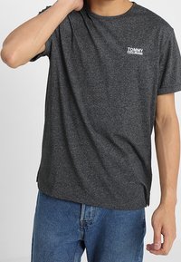 Mörkgrå bomulls T-shirt med rundad halsringning, korta ärmar och en vit "TOMMY JEANS"-logotyp på vänster bröst.