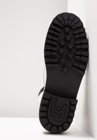 Suela de bota de goma negra con un profundo relieve que presenta patrones de estrellas, marcada con "37" y una impresión de logotipo. Parte superior blanca con un acento de cremallera.