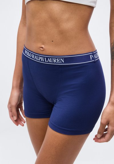Γυναίκα που φοράει μποξεράκια Polo Ralph Lauren σε ναυτικό μπλε με λωρίδα μέσης με λογότυπο, εμφανίζοντας την κοιλιά και μέρος των χεριών της, σε φωτεινό φόντο.
