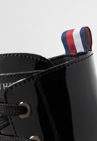 Černá lakovaná kožená bota s kulatou špičkou, látkovými tkaničkami a stříbrnými průchodkami. Má poutko na patě s červenými, bílými a modrými pruhy.