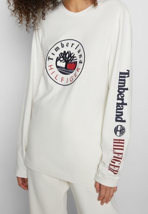 Person wearing white long-sleeve shirt with Timberland and Hilfiger logos on chest and right sleeve, paired with white pants.