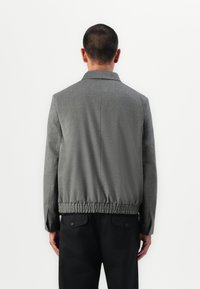 Jachetă gri, texturată, cu guler și tiv elastic; are mâneci lungi și un design curat, minimal. Pantaloni negri vizibili dedesubt.