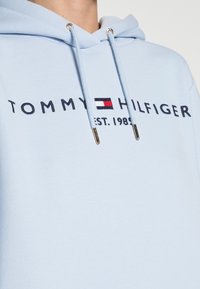 Lichtblauwe hoodie van zachte stof, voorzien van een trekkoordkap en geborduurd "TOMMY HILFIGER EST. 1985" logo in donkerblauw.