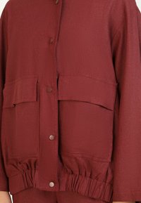 Veste bordeaux en tissu texturé, avec une fermeture frontale à boutons ronds et deux grandes poches poitrine. Détail de taille élastiquée.