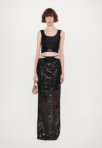 Black cropped tank top with wide straps, paired with a sheer black sequined skirt featuring draped texture and side cutouts. Model holding a patterned bag.