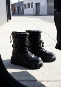 Paire de bottes à plateforme noires épaisses avec lacets posées sur un trottoir urbain ensoleillé à côté d'une portière de voiture ouverte.