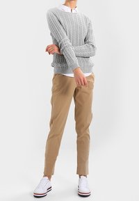 Lichtgrijze kabelgebreide trui met geribbelde manchetten, gecombineerd met beige broek en witte sneakers met gekleurde accenten.