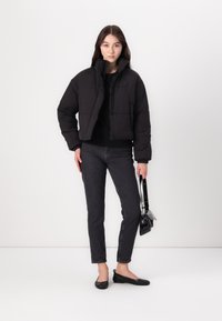 Skraćena crna puffer jakna s visokim ovratnikom, crni pulover, tamnosivi traperice i crne ravne cipele. Model drži malu crnu torbicu.