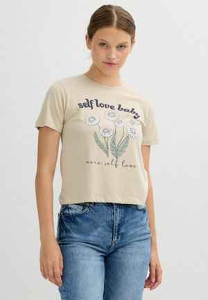 Joven mujer con camiseta beige con flores de margarita y texto "amor propio bebé, más amor propio", combinada con jeans azules, posando contra un fondo blanco.