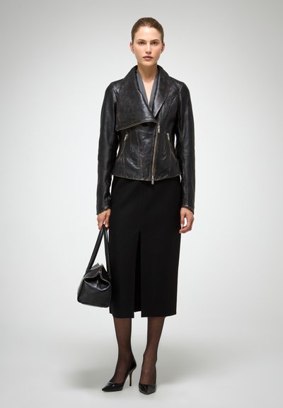 Chaqueta de cuero negra con un cuello caído, dos bolsillos frontales con cremallera, combinada con una falda negra hasta la rodilla, medias transparentes y tacones altos negros.