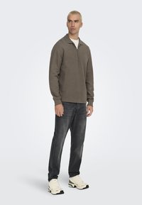 Bruine langemouw pullover met een ritssluiting bij de kraag, gecombineerd met donkergrijze jeans en beige sneakers met groene accenten. Soepele textuur.