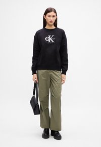 Čierna mikina s logom Calvin Klein, doplnená o olivovo zelené voľné nohavice a čierne platformové čižmy, drží čiernu tašku.