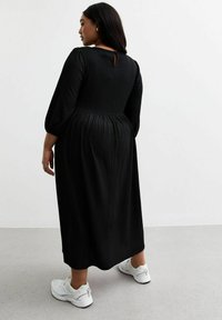 Robe longue noire avec des manches bouffantes et un détail de dos en forme de clé, fabriquée en tissu texturé, associée à des baskets blanches.