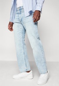 Jeans de mezclilla en azul claro con un diseño de pierna recta, que presentan un ligero desgaste y un dobladillo sin rematar. Combinados con zapatillas blancas.