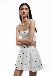 Biała sukienka w czarne kropki, dopasowana góra, falbaniasty dół, cienkie ramiączka związane na ramionach. Modelka ozdobiona błyszczącymi kolczykami.