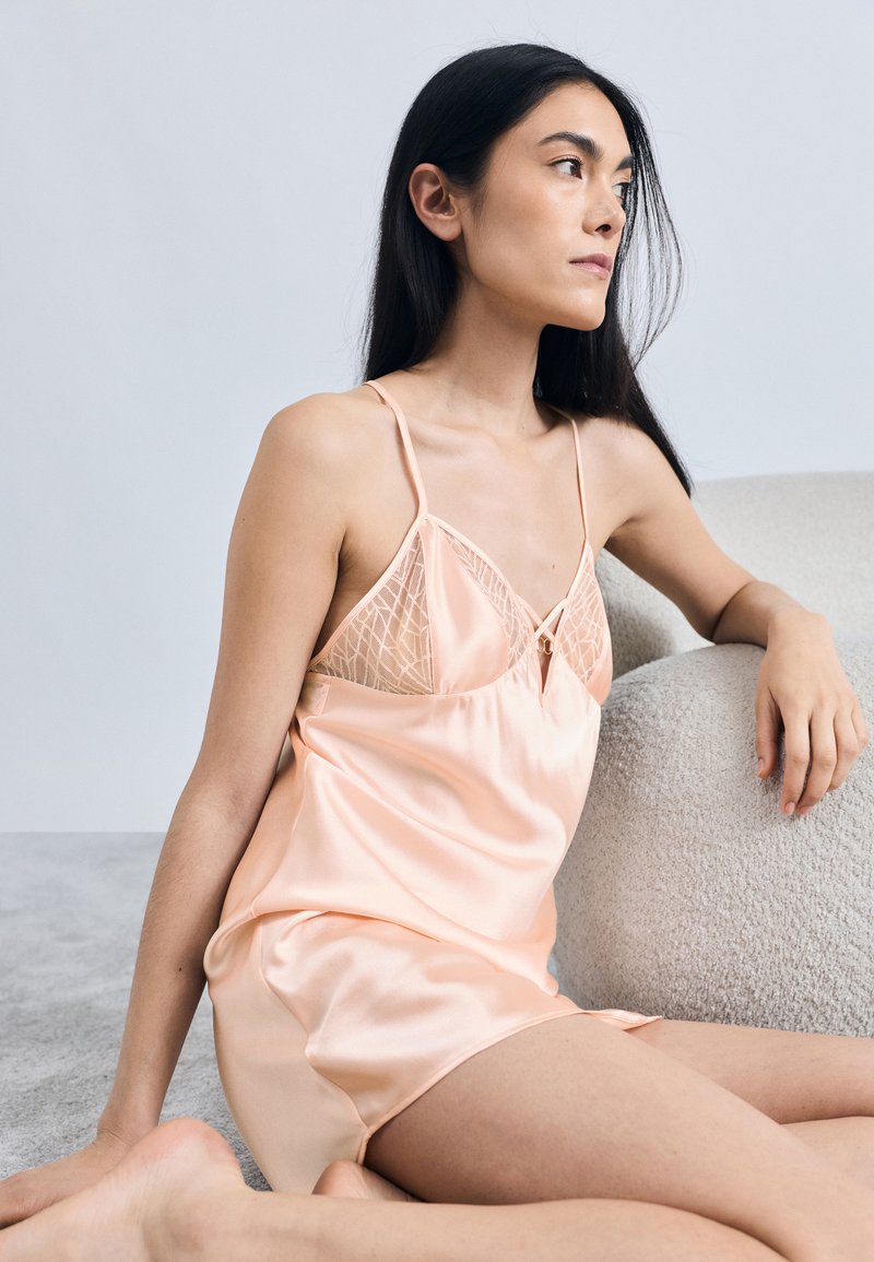 Νυχτικό από σατέν ροδακινί με δαντελένιες λεπτομέρειες, ρυθμιζόμενους ιμάντες και εφαρμοστό μπούστο. Λείο ύφασμα και μήκος μέχρι τα γόνατα.