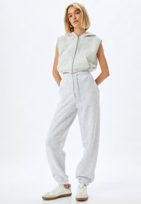 Grijze mouwloze hoodie met rits en een voorzak, gecombineerd met grijze taps toelopende sweatpants. Witte sneakers met tan accenten maken de look compleet.