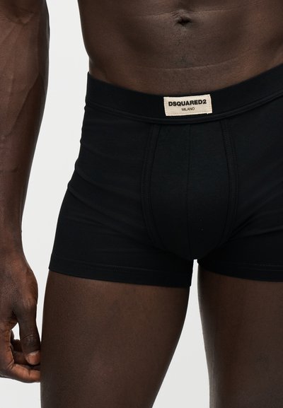 Gros plan d'une personne portant un boxer noir avec une étiquette DSQUARED2 Milano à la taille, montrant le bas du torse et le haut des cuisses.