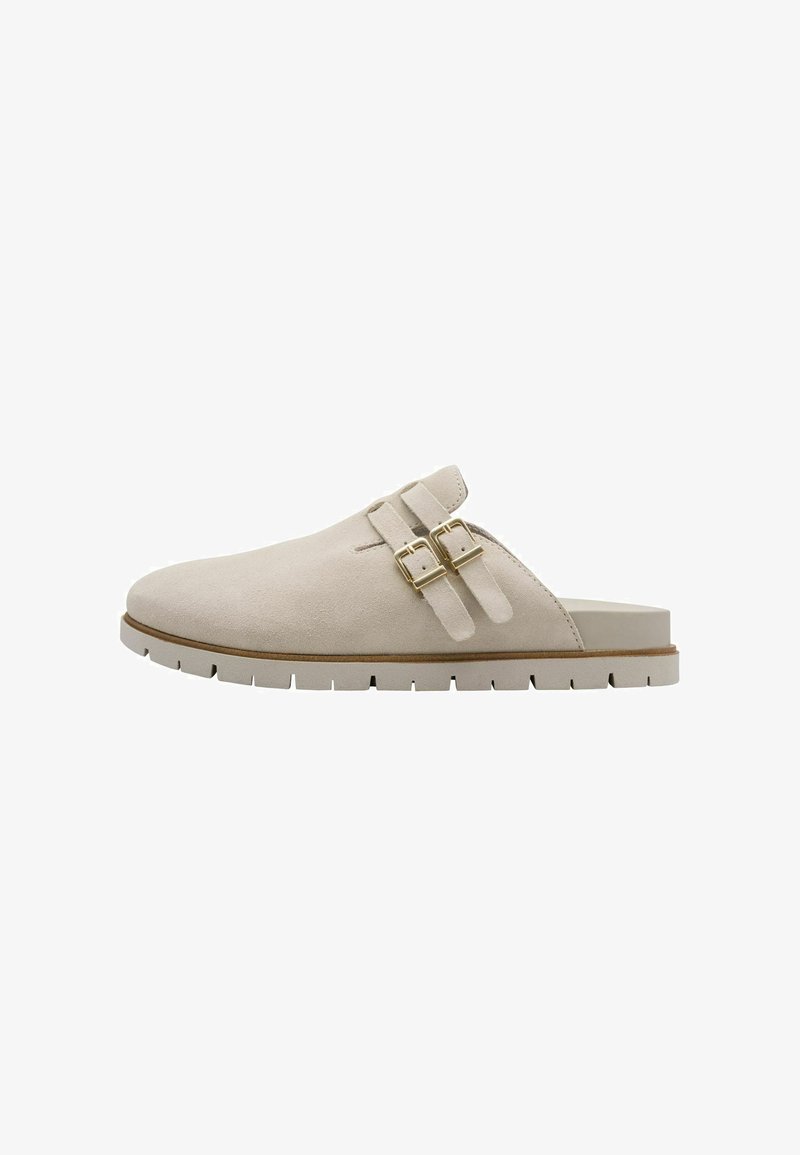 Sabot en daim beige avec deux accents de boucle en or, une épaisse semelle blanche et un bout arrondi. Conçu pour une utilisation facile à enfiler.