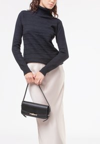 Maglione nero a coste con colletto alto abbinato a una gonna di satin color crema. Il modello tiene una borsa a tracolla in pelle nera con dettagli in oro.