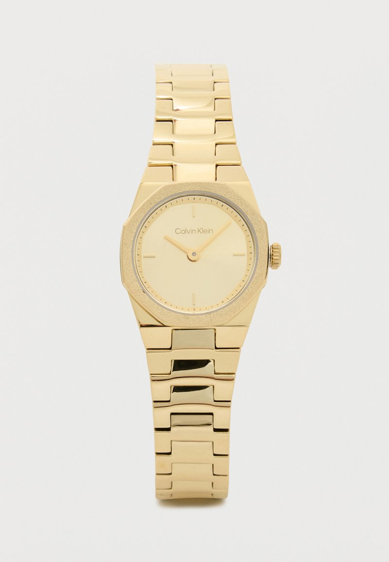 Zelta krāsas rokas pulkstenis ar astoņstūra seju, minimālistisku ciparnīcu un strukturētām detaļām. Pārī savienojamo saišu dizains drošai piestiprināšanai. Zīmola nosaukums redzams.