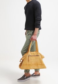 Gul läderväska med två fasta handtag, triangulär form, framficka med dragkedja och mjuk textur, hålls av en person klädd i avslappnad stil.