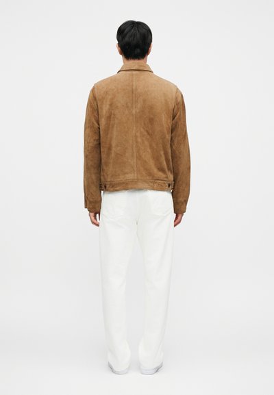 Καφέ σουέτ μπουφάν με γιακά και πίσω καβάλο, συνδυασμένο με λευκό, άνετο παντελόνι. Ελάχιστα ορατά μεταλλικά στοιχεία και διακριτική σήμανση.