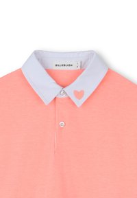Polo shirt rose pâle avec un col bleu clair, présentant un cœur brodé sur le côté gauche du col et trois boutons au niveau du cou.