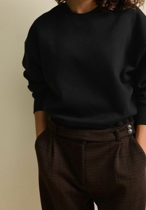 Personne portant un haut noir à manches longues rentré dans un pantalon marron à carreaux taille haute avec des boutons argentés, mains dans les poches.