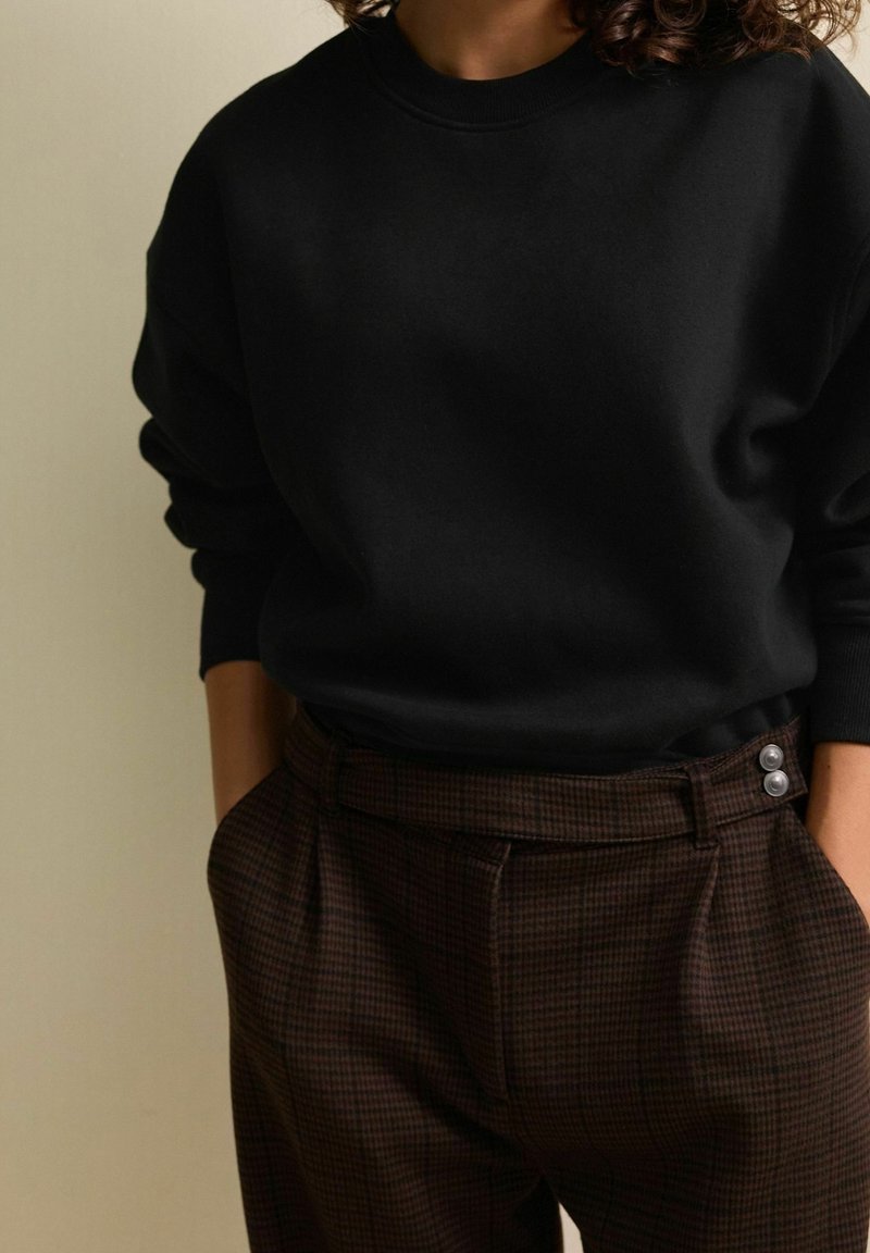Personne portant un haut noir à manches longues rentré dans un pantalon marron à carreaux taille haute avec des boutons argentés, mains dans les poches.