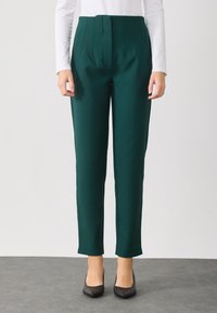 Groene op maat gemaakte broeken met een gladde textuur en gestructureerde pasvorm. Bevat plooien bij de taille en een taps toelopend ontwerp. Een paar zwarte schoenen.