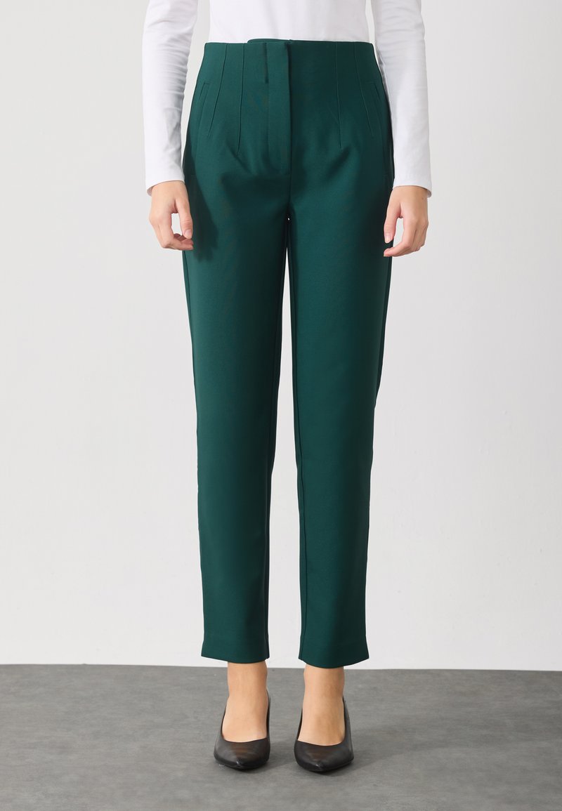 Groene op maat gemaakte broeken met een gladde textuur en gestructureerde pasvorm. Bevat plooien bij de taille en een taps toelopend ontwerp. Een paar zwarte schoenen.