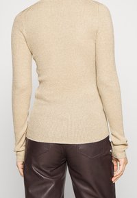 Pull côtelé beige avec une coupe ajustée, manches longues et un léger éclat subtil. Associé à un pantalon en cuir marron, mettant en valeur le contraste des textures.