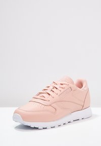 Ljusrosa träningsskor med en slät läderöverdel, perforerad detaljering och en vit gummisula. Rund tå och snörningsdesign.